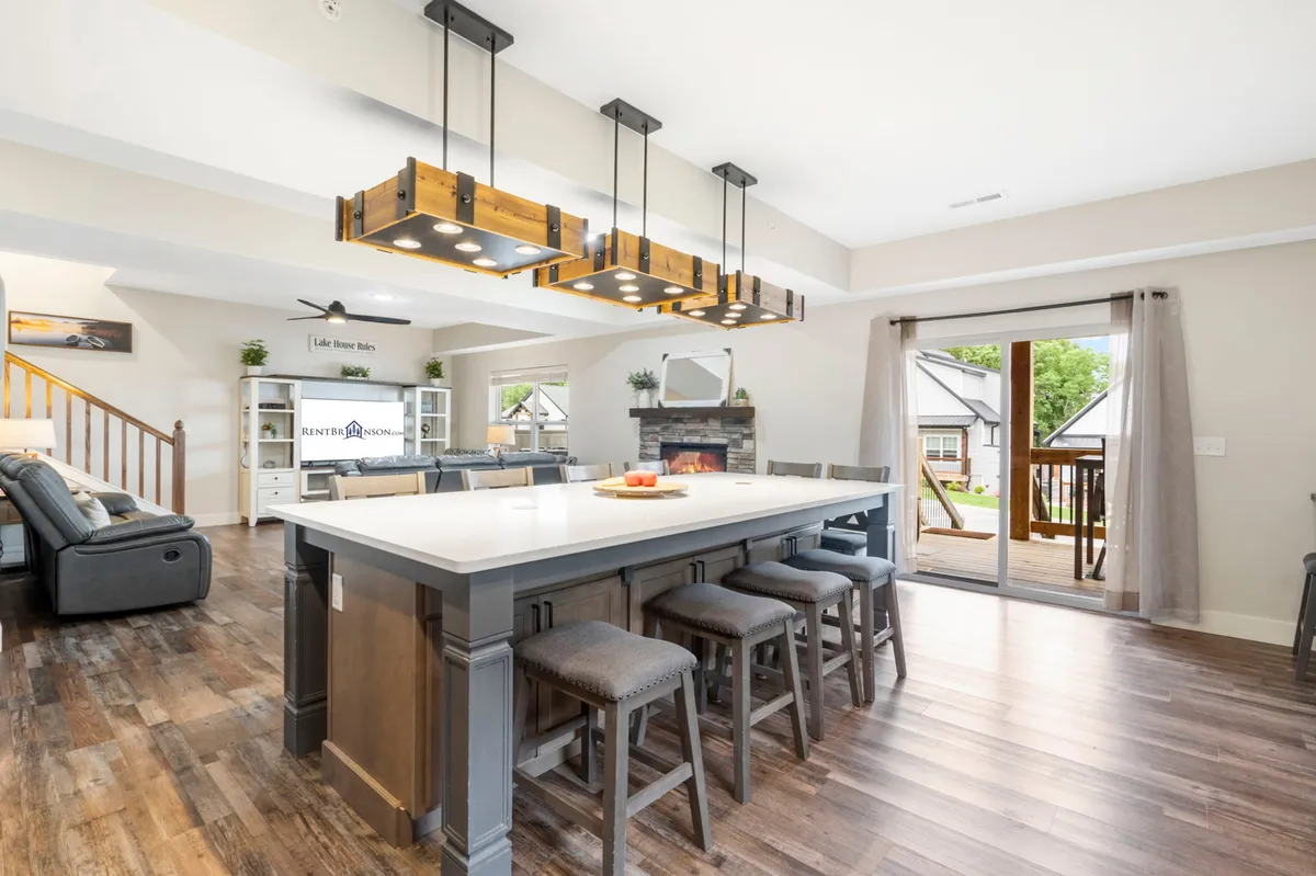 A luminous open-concept living space with a kitchen island, contemporary lighting fixtures, a cozy fireplace, and a sliding door that opens directly to the deck. The area flows seamlessly from kitchen to dining and living spaces, with ample room for family gatherings and entertaining guests.