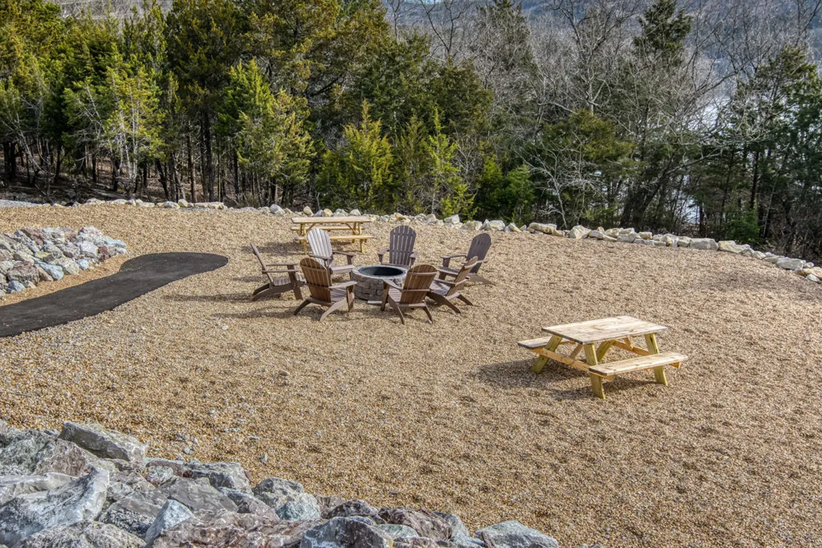 A cozy outdoor fire pit surrounded by Adirondack chairs and a picnic table, set against a backdrop of natural stone and greenery. The setting evokes evenings spent socializing, stargazing, and enjoying the outdoors in comfort.