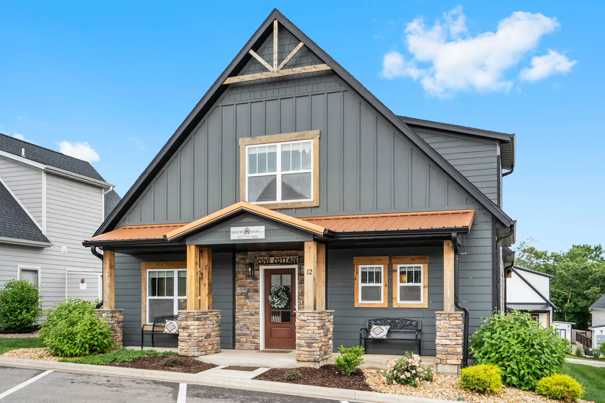 A welcoming view of a wooden porch accented with stone, large windows, and a sturdy metal roof. The porch is set against a backdrop of greenery, conveying a sense of privacy and connection to nature. Ample space is available for gatherings or quiet relaxation, and the architectural details hint at upscale comfort.