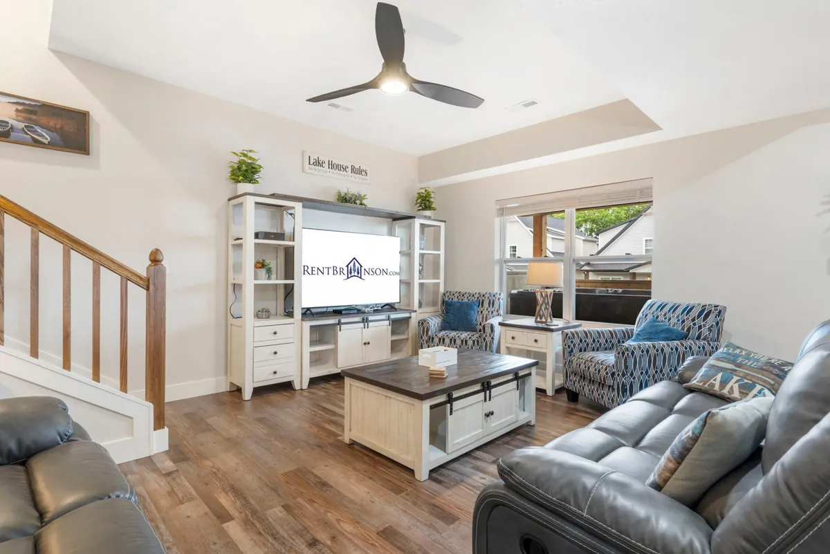 A spacious, comfortable living room featuring plush sofa seating, a TV unit accented with decorative items, and a staircase leading to the upper floor. The room exudes a casual, welcoming vibe ideal for movie nights or unwinding after a day at the lake.