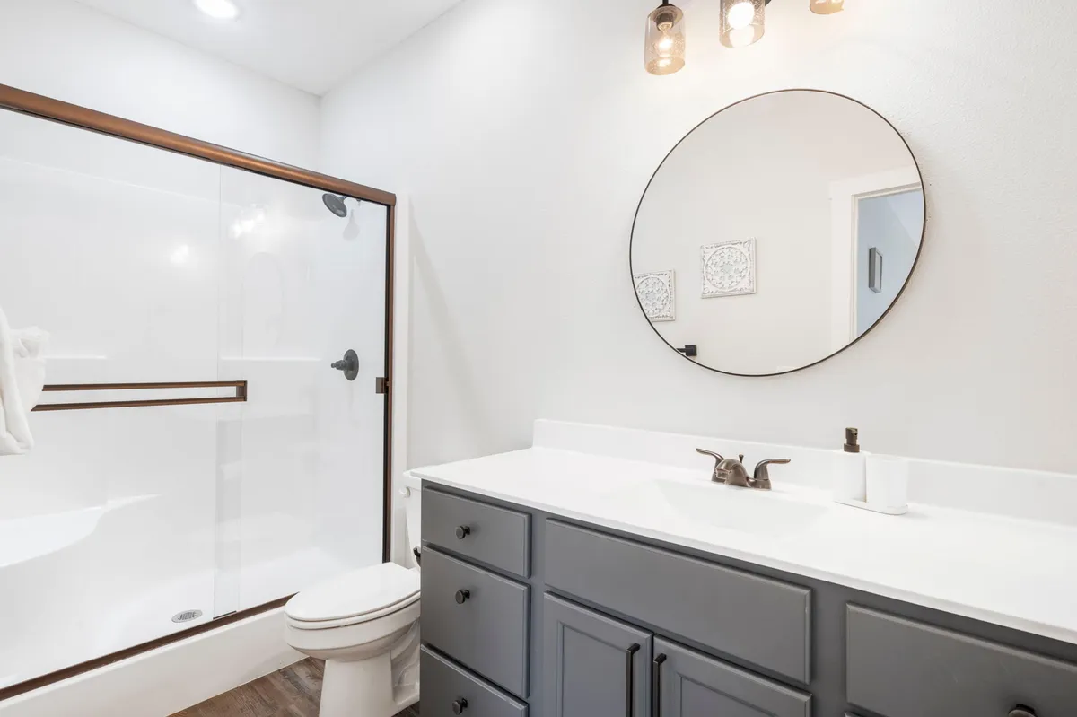 A modern, inviting bathroom with a stylish vanity featuring dark cabinets, a large round mirror, and a glass-door shower. The space is bright and designed for comfort, perfect for refreshing and unwinding.