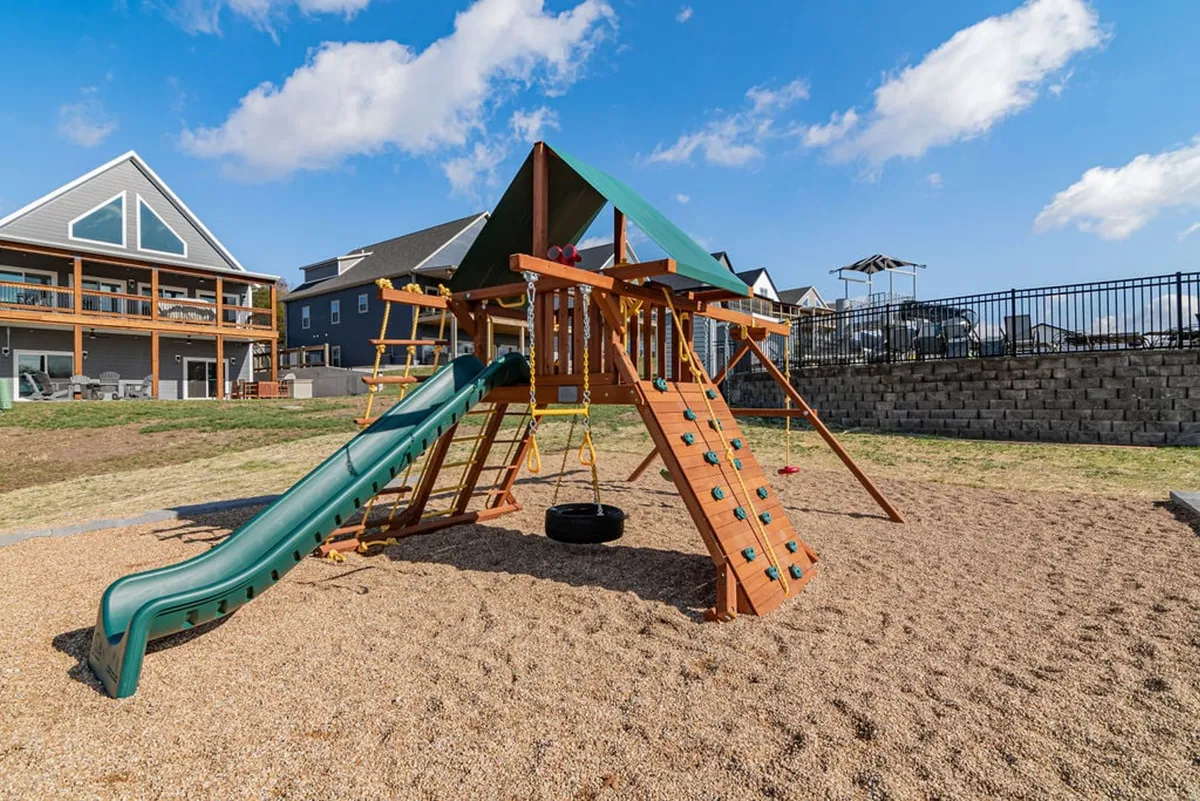 A lively playground scene featuring a large play structure, slide, and swing set bathed in sunshine. The space is surrounded by greenery and set within a family-friendly neighborhood, inviting children and families to enjoy outdoor play and social gatherings.